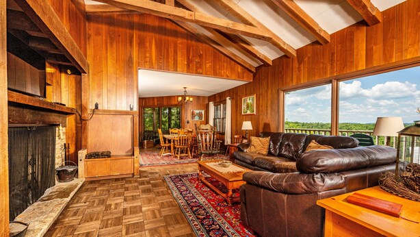 Sala de estar com piso de madeira, lareira, sofá de couro e uma grande janela com vista para o verde.