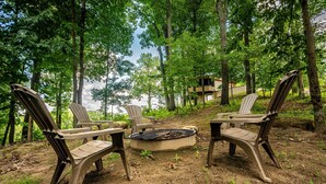 Um camping com cadeiras Adirondack ao redor de um braseiro, cercado por árvores e vista para um lago.