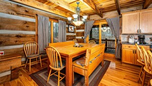 L’intérieur d’un chalet en bois comprend une table à manger, des chaises, un banc, un coin cuisine et des rideaux à carreaux.