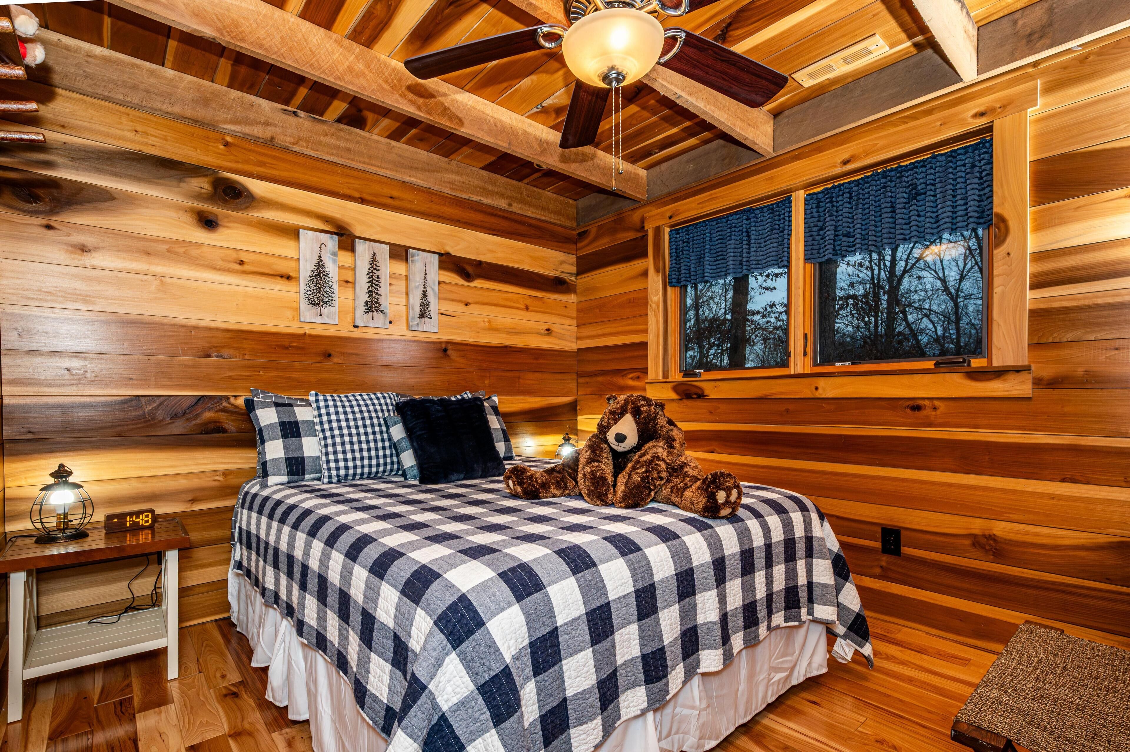 Ein Schlafzimmer in einer Holzhütte mit einem Bett, einem Nachttisch, einem Teddybären und einem Fenster mit Blick auf Bäume.