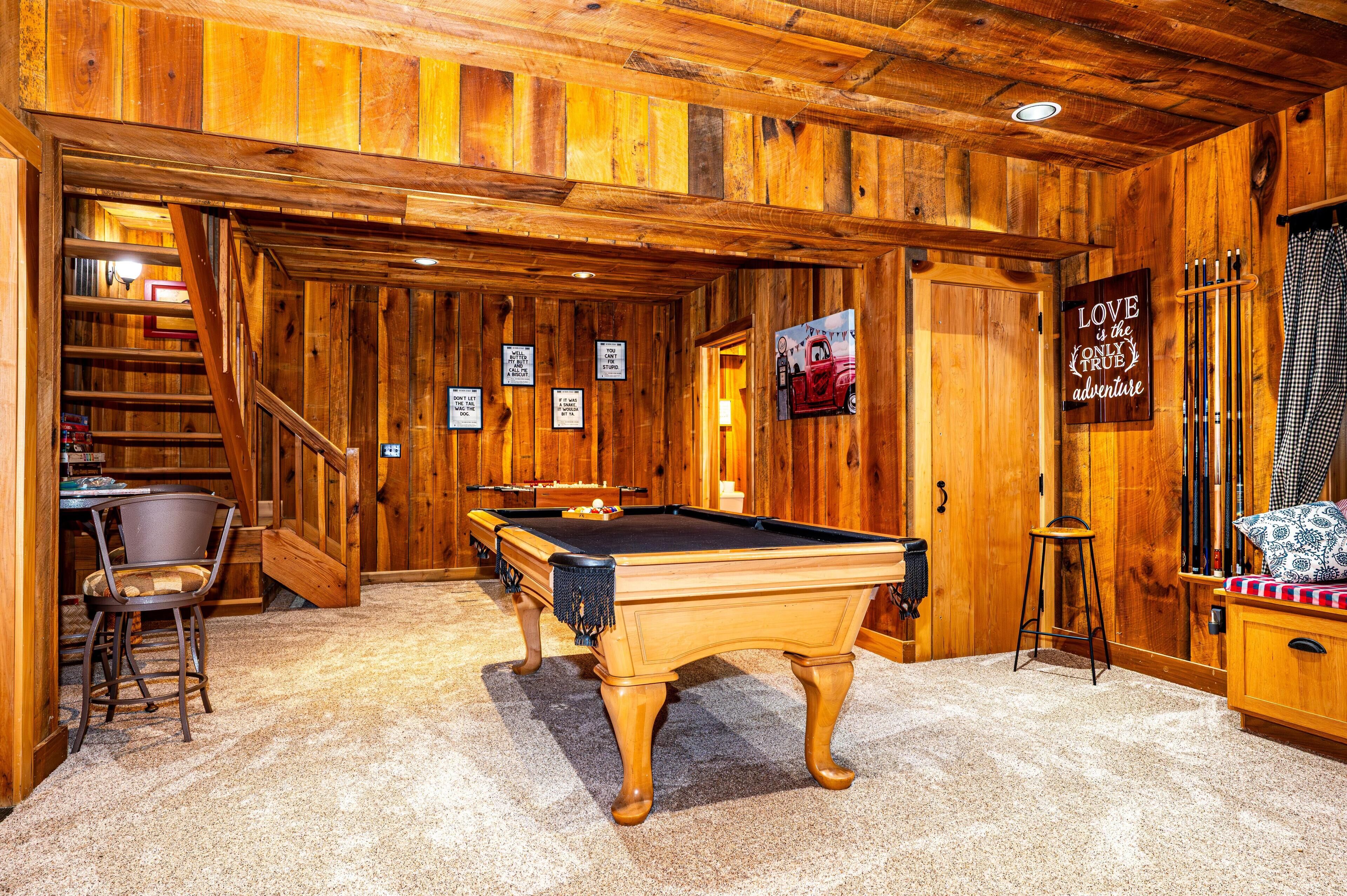 Une table de billard en bois, dans une pièce où se trouvent un escalier, une chaise de table de billard et un petit tabouret.