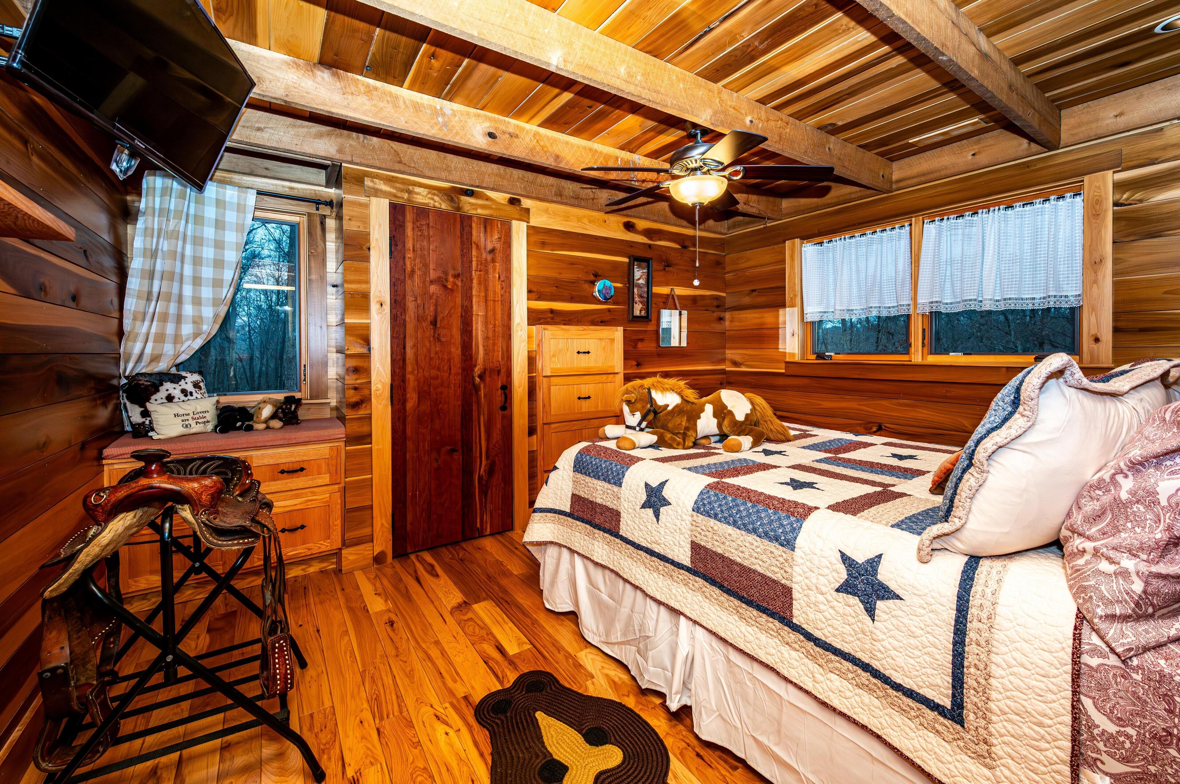 Dormitorio de cabaña de madera con cama, ventilador de techo, ventana con cortinas y cómoda de madera.