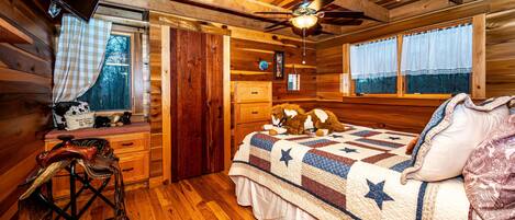 Dormitorio de cabaña de madera con cama, ventilador de techo, ventana con cortinas y cómoda de madera.