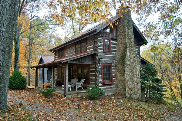 Log cabin exterior with antique wood, red-trimmed windows, stone chimney, and covered porch with wicker furniture.