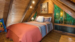 Una camera da letto mansardata accogliente, con un soffitto spiovente, una grande finestra vista su del verde, un letto con biancheria a motivi decorativi e un quadro appeso al muro.