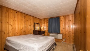 Habitació amb paret revestida de fusta, un llit amb matalàs, una commoda amb mirall, una làmpada i una finestra amb cortina blava.