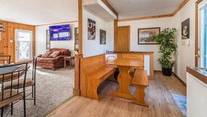Ein gemütliches Zimmer mit einer Holzbank, einem Esstisch und einer Couch. An den Wänden hängen Bilder in Rahmen und es steht ein Blumentopf bereit.