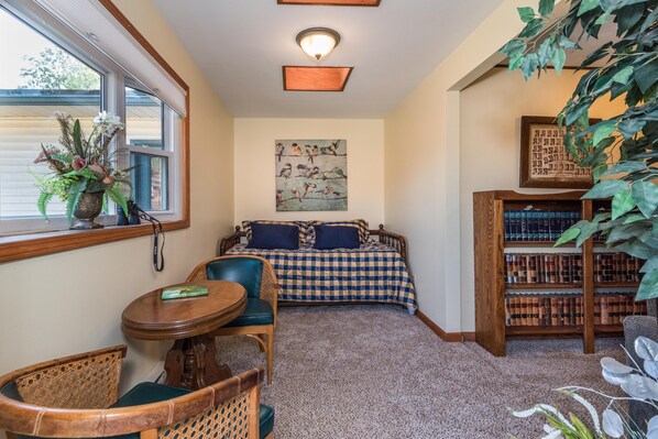 A cozy room with a checkered bed, a wooden bookshelf, a round table, and a large window with a view.