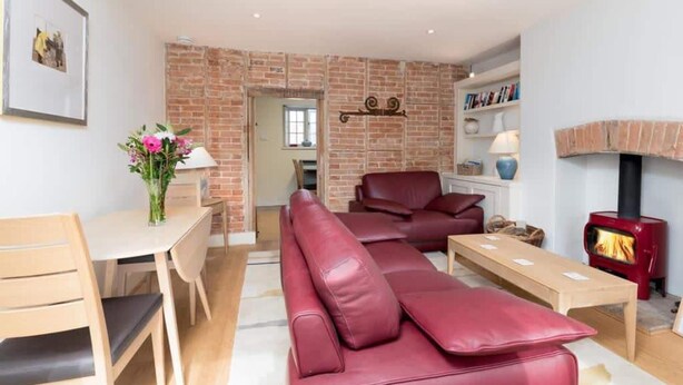 A living room with a brick wall, a sofa, a table, a fireplace, and a bookshelf. - Stunning Driftwood Cottage at Holme-next-the-sea (Hunstanton)