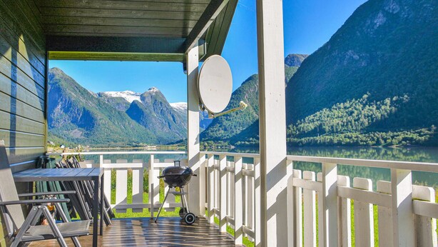 Um deck de madeira com churrasqueira, guarda-corpo branco e vista para um lago e montanhas.