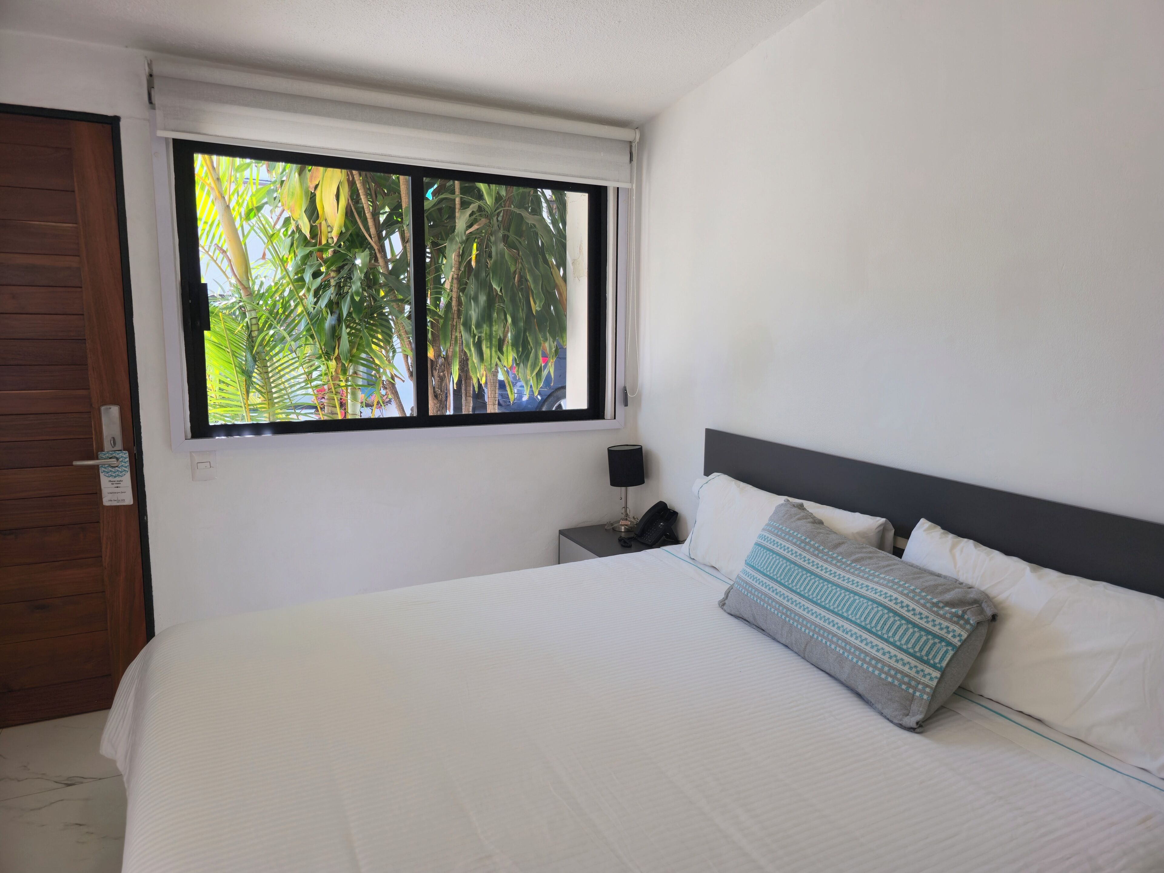 Een slaapkamer met een groot bed, nachtkastje, telefoon en een raam met uitzicht op palmbomen.