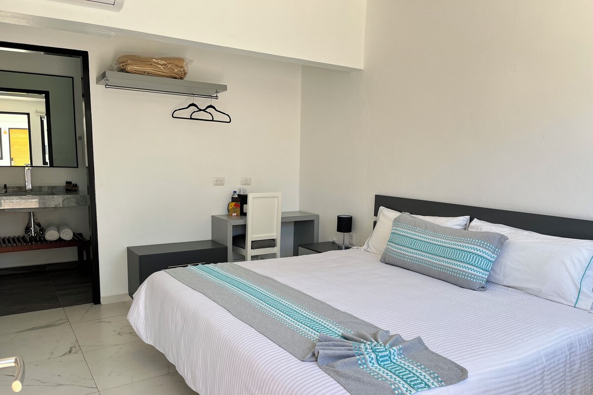 Ein modernes Hotelzimmer mit einem ordentlich bezogenen Bett, einem Nachttisch mit Lampe, einem kleinen Schreibtisch und einem durch eine geöffnete Tür einsehbaren Badezimmer.