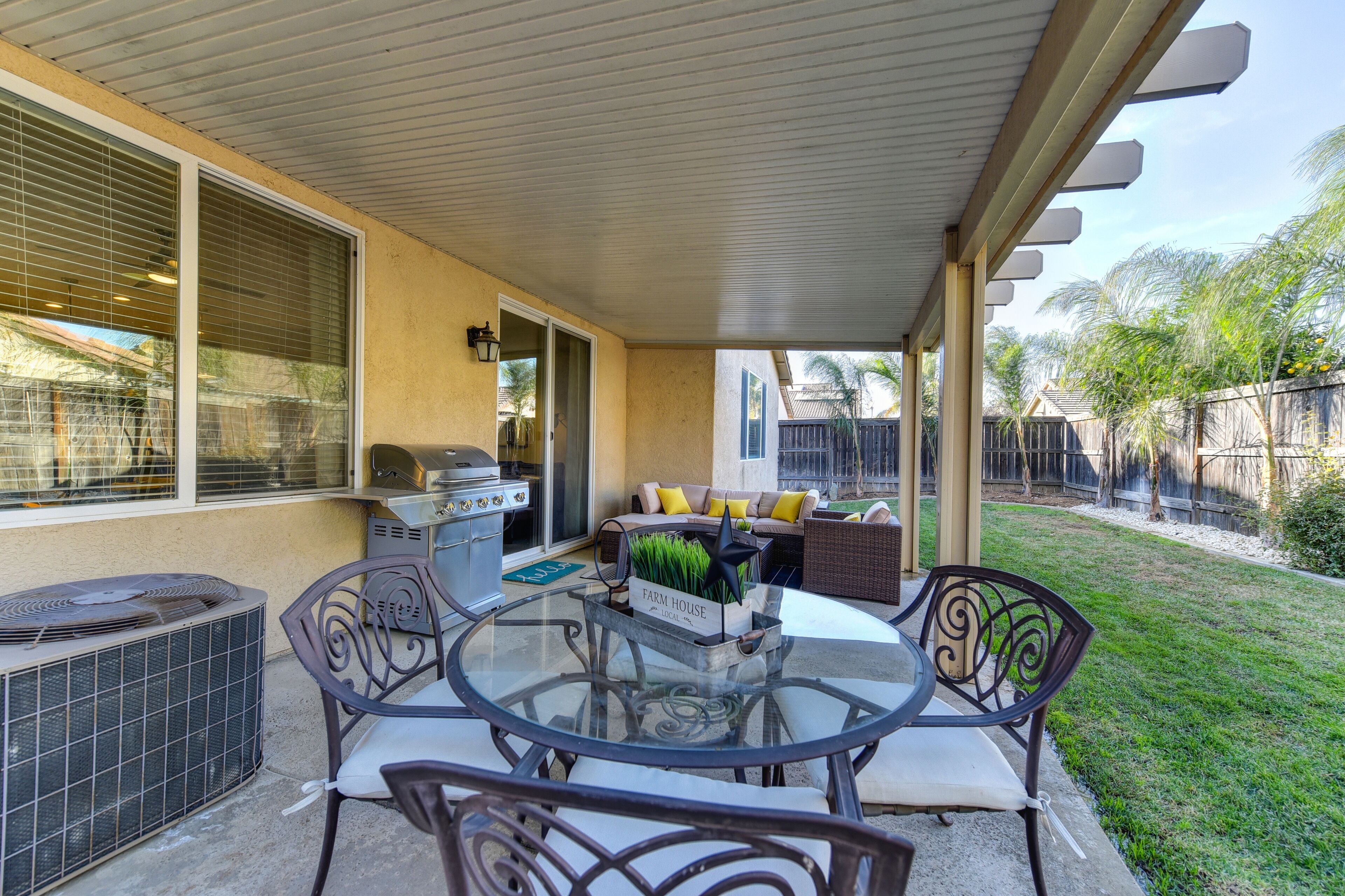 An outdoor patio with a glass table, metal chairs, a grill, and a covered area with a view of the backyard.