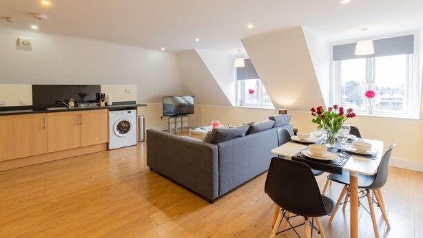 A modern kitchen with a washing machine, a grey sofa, and a dining area with a table set for two. - 2-bedroom Penthouse, Clockhouse, Hoddesdon (Hoddesdon)
