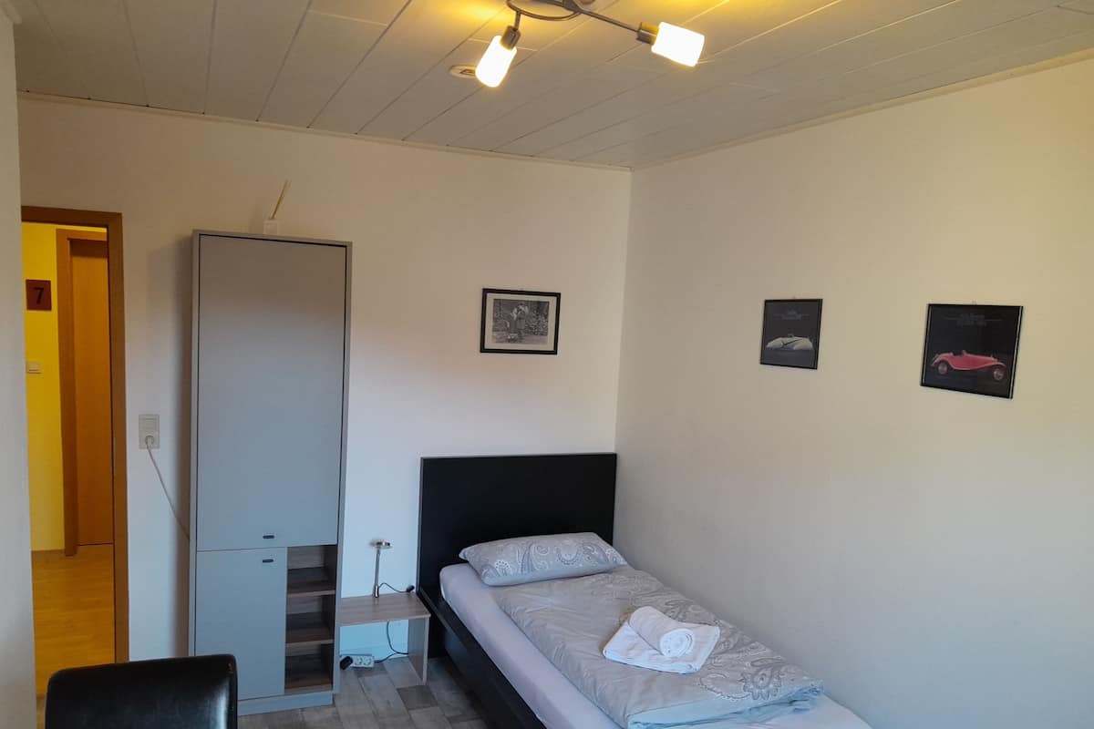 A small, neatly arranged bedroom with a single bed, a chair, and a desk. There are three wall-mounted lights, a door, and a few framed pictures.