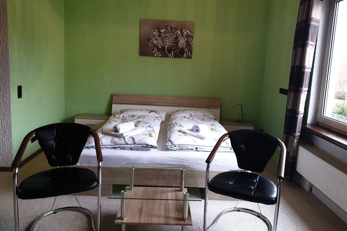 A bedroom with a bed, two chairs, a table, a zebra artwork, and a window with curtains.