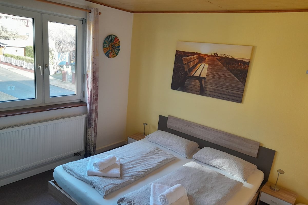 A hotel room with a bed, a window with curtains, a wall clock, and a painting.