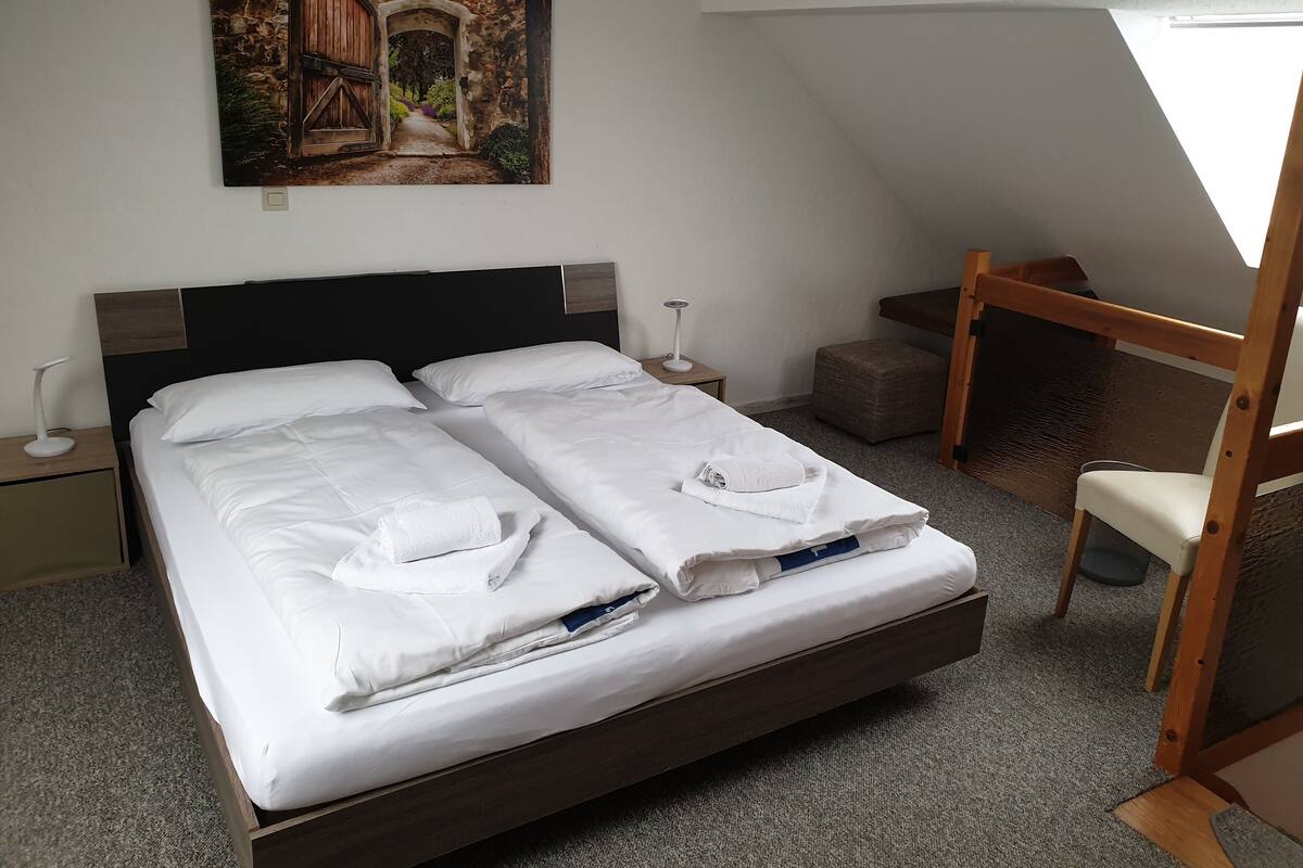 A double bed with white linens, a wooden headboard, a nightstand with a lamp, a painting of a stone archway, a small wooden desk, and a chair.