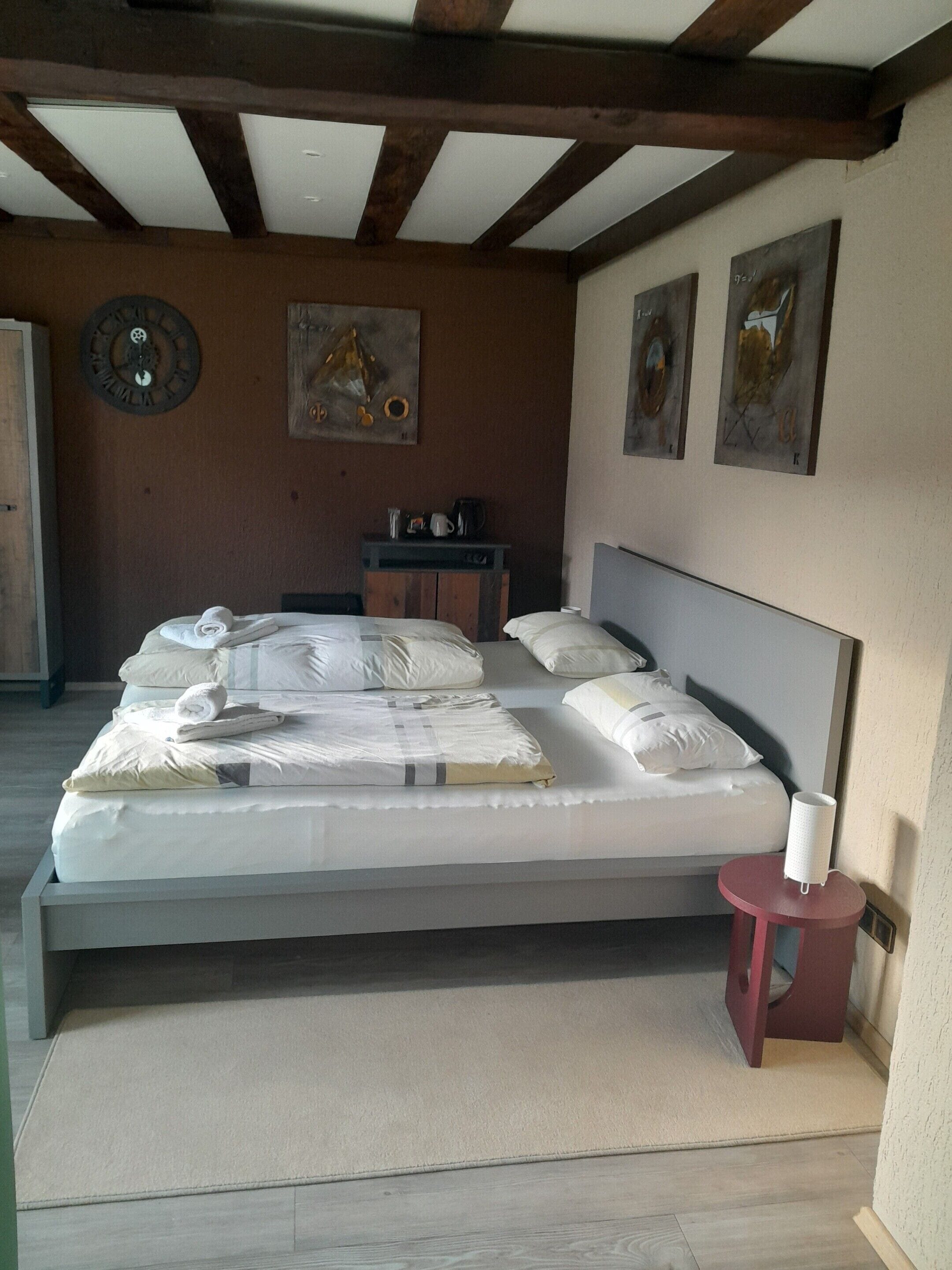A neatly made bed with pillows and a rolled towel, a small red table with a white object, and artwork on the walls.