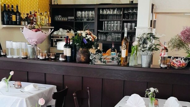 A restaurant interior with tables set for dining, a bar area with shelves of bottles and glasses, and a yellow textured wall. - Pension Zum Adler (Hünstetten)