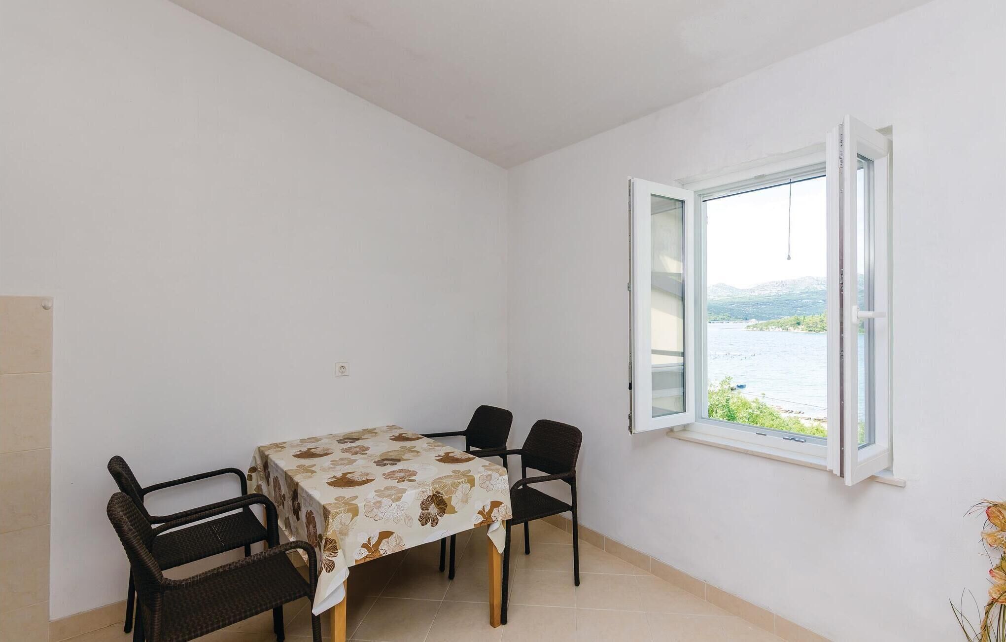 Un comedor con una mesa cubierta con mantel floral, cuatro sillas y una ventana con persiana entreabierta.