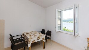 A dining area with a table covered in a floral tablecloth, four chairs, and a window with an open shutter. - Stunning apartment in Hodilje with house sea view (Hodilje)