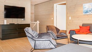 Ein modernes Wohnzimmer mit einem an der Wand befestigten Flachbildfernseher, einer Holzkommode, einem grauen Sofa, einem braunen Ledersessel und einem runden Tisch mit einem orangefarbenen Kissen.