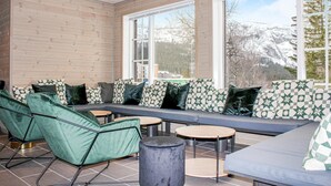 Ein modernes Interieur mit einer Holzwand, einem großen Fenster mit Blick auf einen verschneiten Berg und einem grünen Sofa mit gemusterten Kissen sowie einem runden Tisch.
