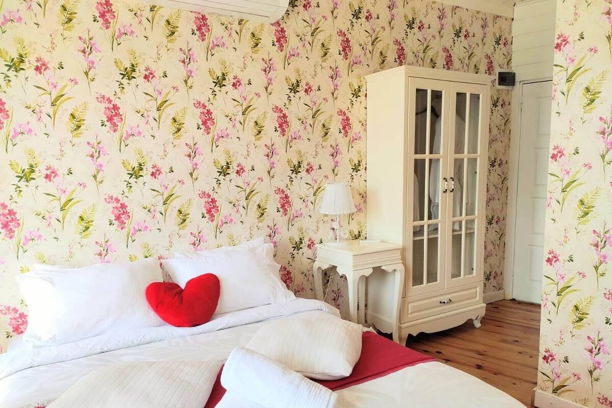 A bedroom with floral wallpaper, a bed with white bedding and red pillows, a white cabinet, and a nightstand with a lamp.