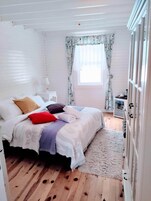 Un dormitorio con cama, mesita de noche, ventana con cortinas y suelo de madera.