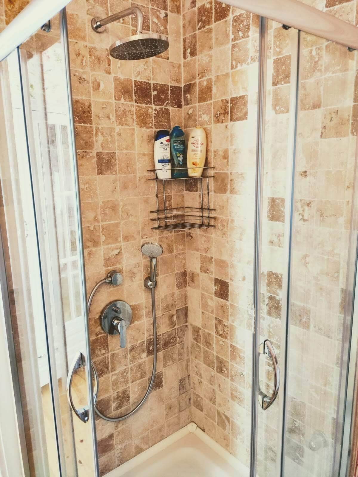 A glass-enclosed shower with a tiled wall, a handheld showerhead, a wall-mounted showerhead, and a shelf with toiletries.