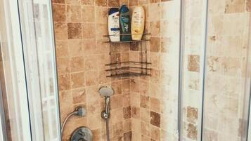 A glass-enclosed shower with a tiled wall, a handheld showerhead, a wall-mounted showerhead, and a shelf with toiletries.