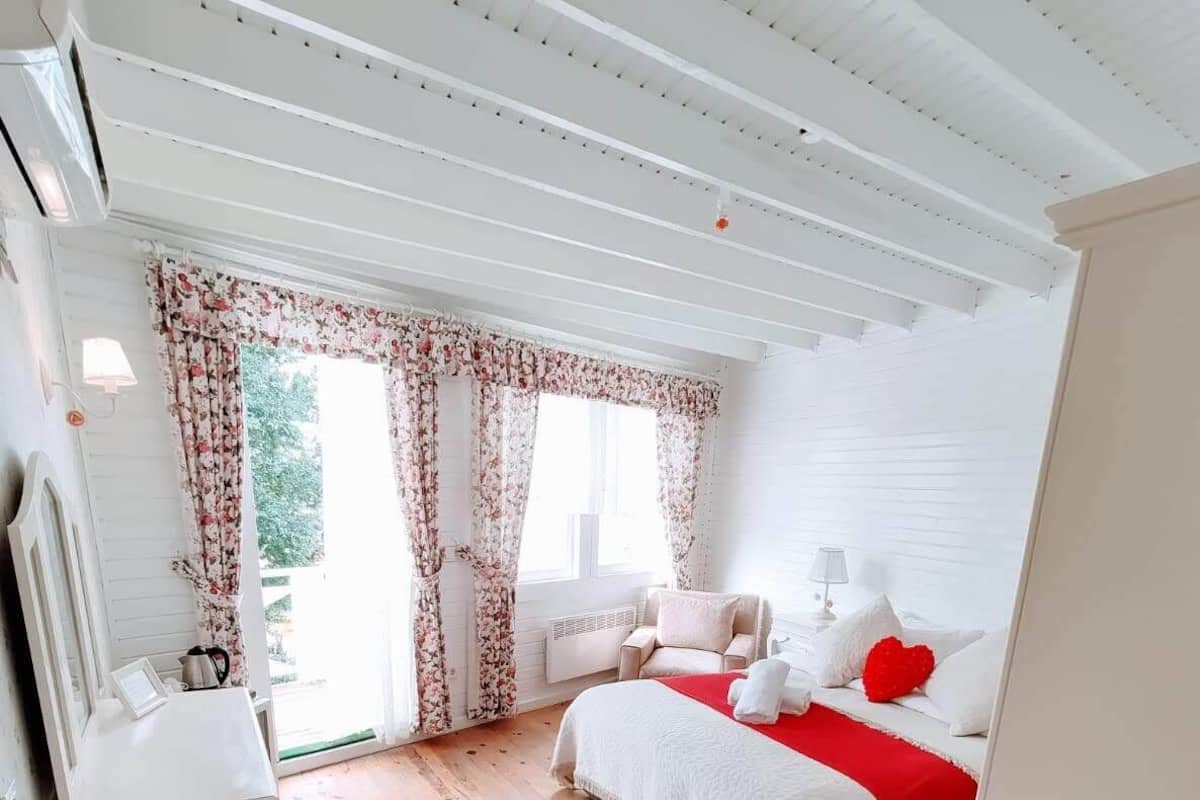 A bedroom with a wooden floor, a bed with white bedding and red accents, a chair, a bedside table, and a window with floral curtains.