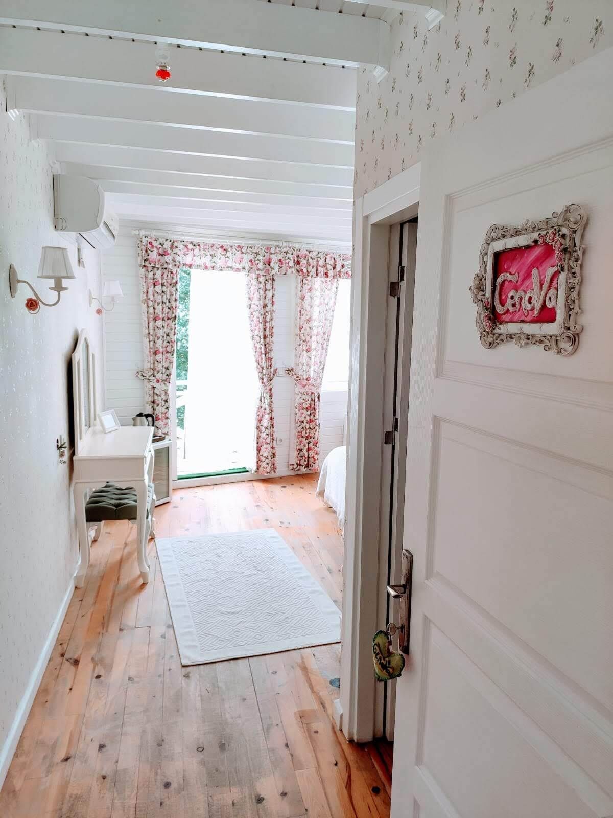 Ein gemütliches Schlafzimmer mit Holzboden, einem Bett mit weißer Bettdecke, einem kleinen Tisch mit Lampe und einem Fenster mit floralen Vorhängen.