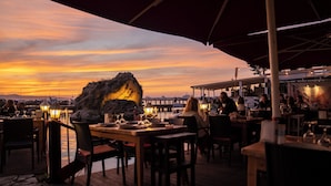 Un espacio de comedor al aire libre con mesas y sillas, con vistas a un cuerpo de agua y un promontorio rocoso durante el atardecer.