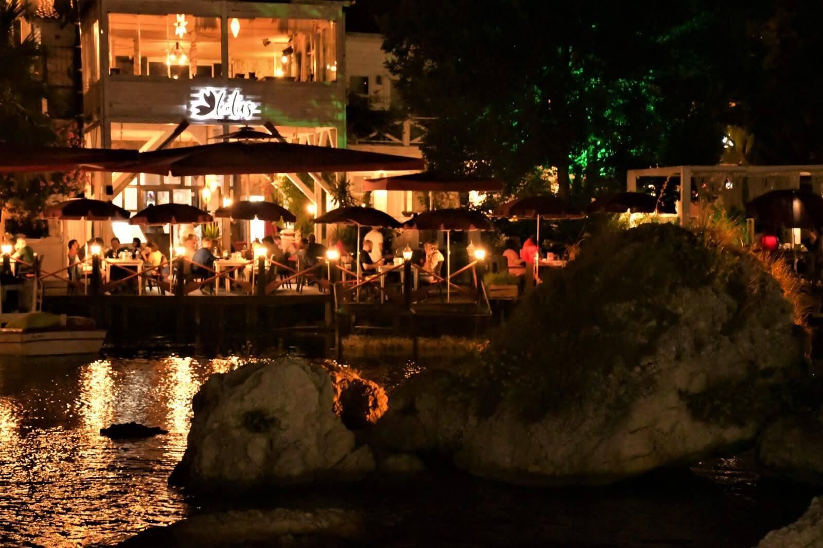 Un área de comedor al aire libre con sombrillas y mesas, una fuente de agua en primer plano y un edificio con señalización iluminada al fondo.