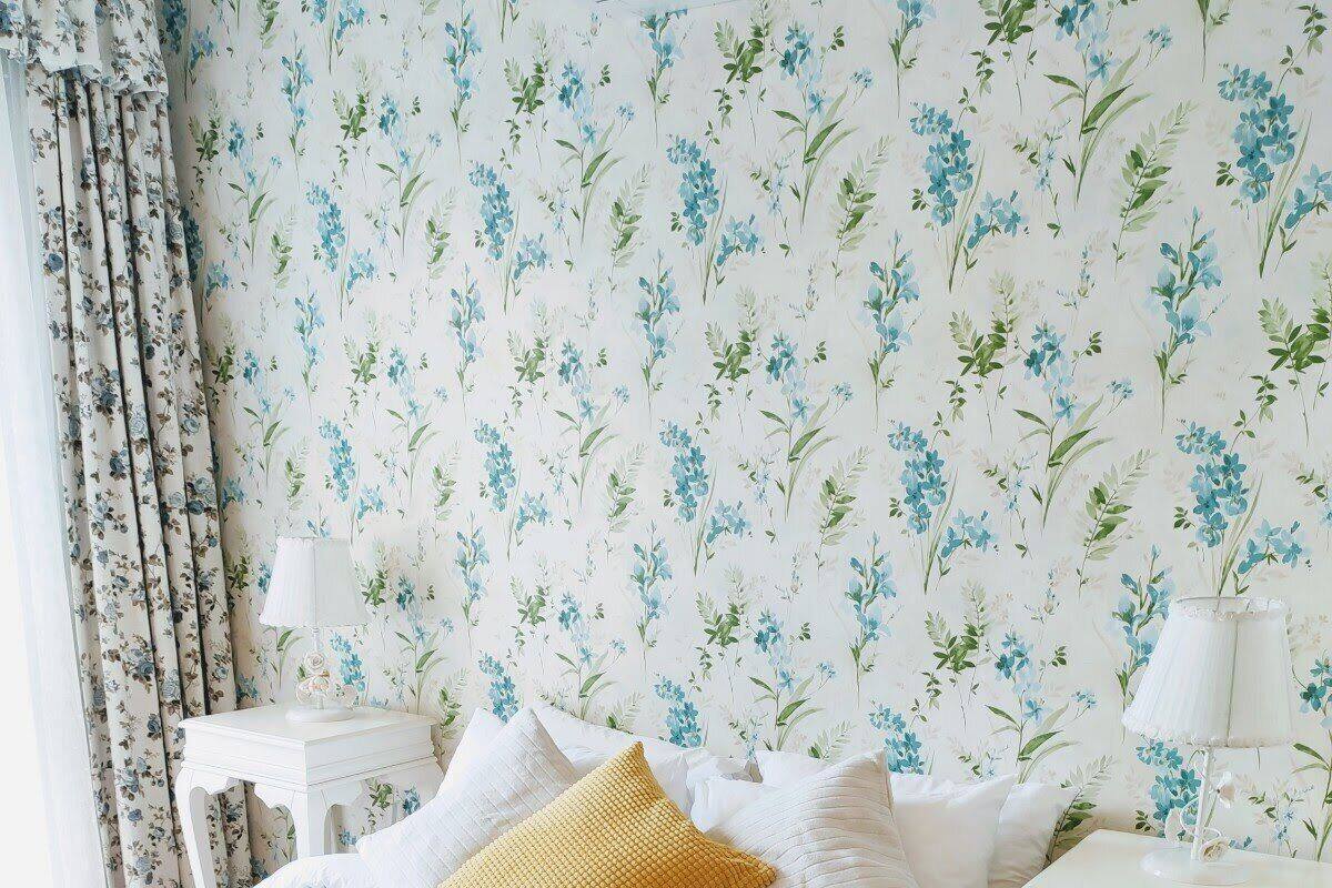 A neatly made bed with white and yellow pillows, a blue blanket, and a rolled white towel. There are two bedside tables with lamps and a floral wallpaper background.