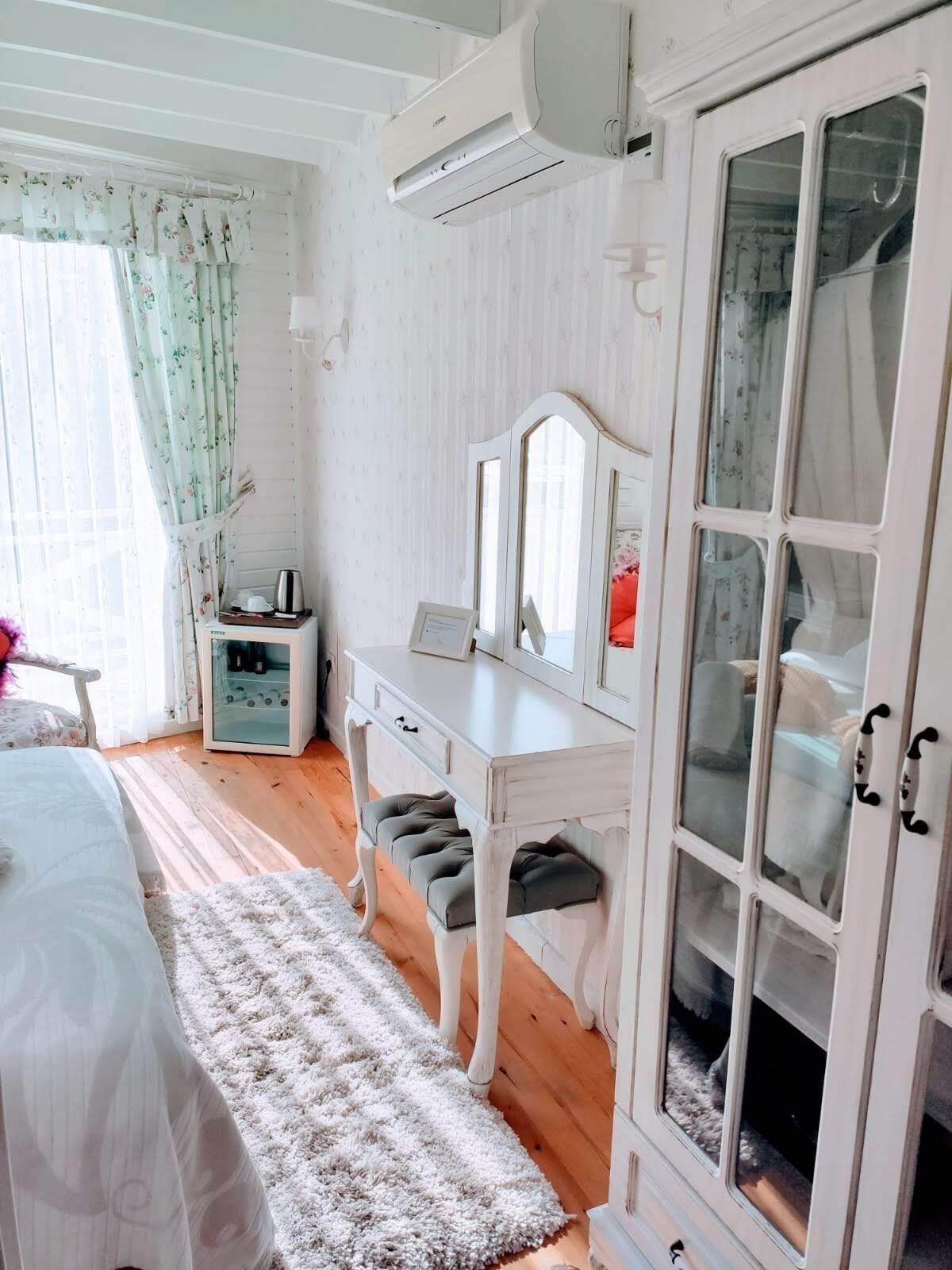 A bedroom with a bed, a dresser, a vanity with mirrors, a small cabinet, and a fluffy rug.
