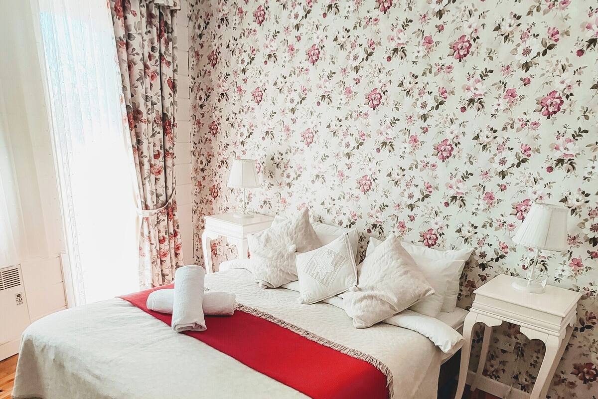 A bedroom with a bed, a nightstand, a red throw blanket, floral wallpaper, and wooden flooring.