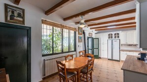 A dining area with a wooden table and chairs, a window with a view of greenery, a ceiling fan, and a kitchen with white cabinets and a refrigerator.
