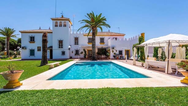 A large white building with a swimming pool in front, surrounded by well-maintained grass and palm trees.
