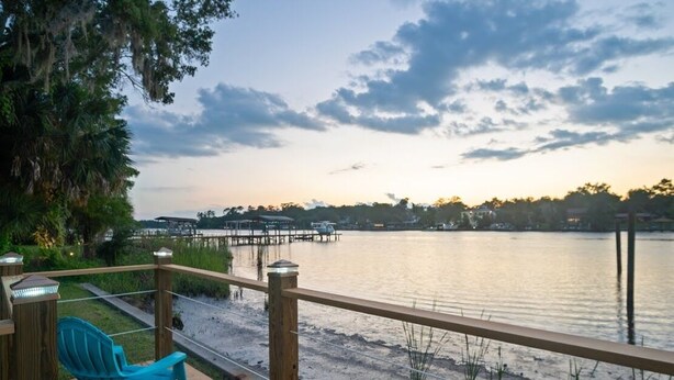 A wooden deck with a railing overlooking a calm lake, a dock extending into the water, and a blue chair on the deck. - Riverfront Retreat. Private Oasis 7 minutes to downtown (Jacksonville)