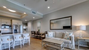 A modern kitchen with white cabinetry, a dining area with a white table and chairs, and a living room with a sofa and a large mirror. - Seaside Bliss with Spa | Luxe 6-Guest Haven (Destin)