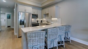 Cocina moderna con armarios blancos, electrodomésticos de acero inoxidable, una isla central con sillas de mimbre y una lámpara de techo luminosa.