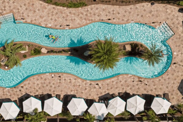 Vue aérienne d’une piscine à la forme ondulée, entourée de palmiers et de chaises longues dotées de parasols.