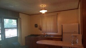 A compact kitchen with a sink, a refrigerator, and a coffee maker. There is a window with curtains, a small table, and a chair. - The Gold Eagle Cabin (Lake George)