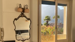 A room with a bed, a desk with a lamp, a radiator, and a window with a view of trees and buildings. - App.with garden, WI-FI free, automatic gate, covered garage, air conditioning  (Bellagio)