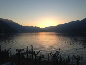 Um pôr do sol sobre um lago calmo, com montanhas ao fundo e um cais com plantas de cactos em primeiro plano.