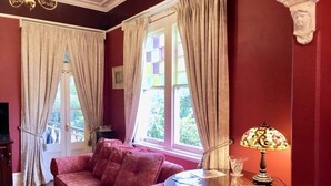 Ein Zimmer mit roter Wand, einem rosafarbenen Sofa, einem runden Holztisch mit Lampe und einem Fenster mit Vorhängen.