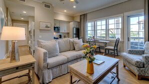 Un salon spacieux avec un canapé, une table basse et une table à manger. On y trouve un ventilateur de plafond, une fenêtre avec vue et, au fond, une cuisine.